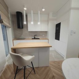Klemba Design - Nowoczesna, jasna kuchnia z drewnianym blatem i wyspą. Białe szafki, czarny okap i sprzęt AGD. Krzesło barowe z beżową tapicerką. Parkiet ułożony w jodełkę.