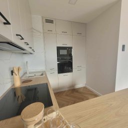 Klemba Design - Jasna kuchnia z drewnianym blatem i podłogą w jodełkę. Białe szafki z czarnymi uchwytami, czarna płyta indukcyjna i sprzęt AGD. Na blacie szklane naczynia.