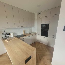 Klemba Design - Jasna kuchnia z drewnianym blatem i parkietem w jodełkę. Minimalistyczne szafki z czarnymi uchwytami. Na blacie akcesoria kuchenne i szklanki na tacy.