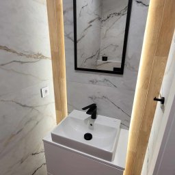 Klemba Design - Nowoczesna łazienka z umywalką nablatową, czarną armaturą i lustrem w czarnej ramie. Marmurowe płytki ścienne i podświetlane drewniane panele.