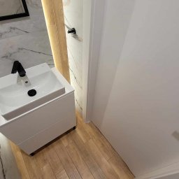 Klemba Design - Nowoczesna łazienka z białą umywalką nablatową i armaturą w kolorze czarnym. Marmurowe płytki ścienne i drewnopodobna podłoga tworzą elegancki kontrast.