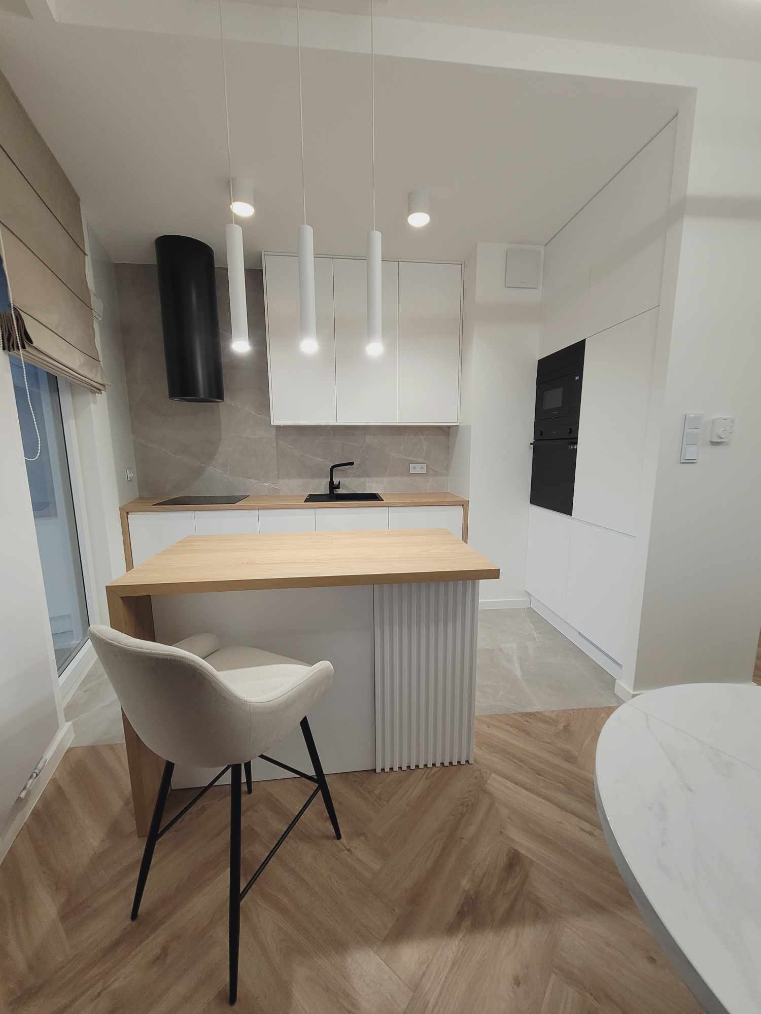 Nowoczesna, jasna kuchnia z drewnianym blatem i wyspą. Białe szafki, czarny okap i sprzęt AGD. Krzesło barowe z beżową tapicerką. Parkiet ułożony w jodełkę.