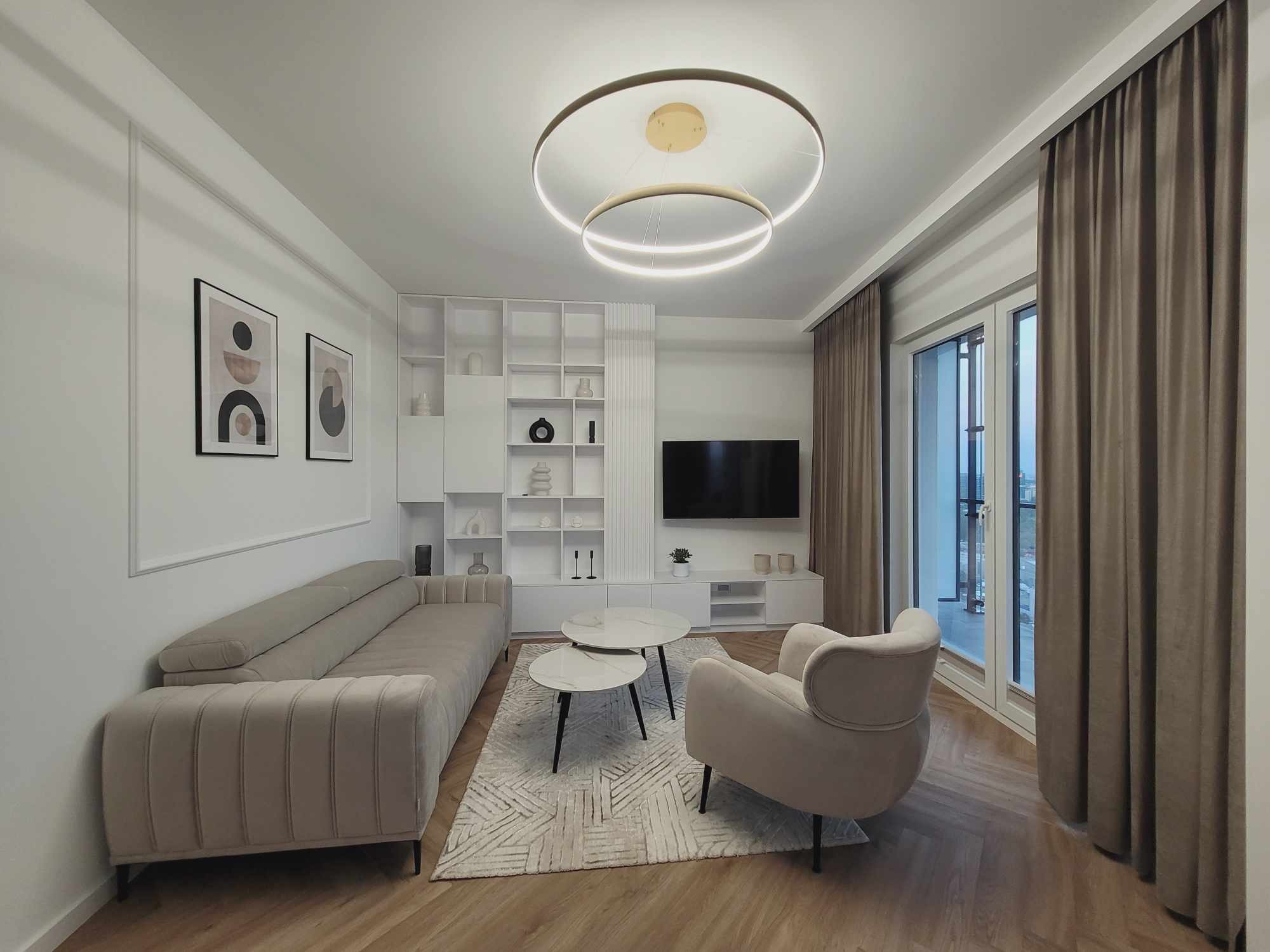 Jasny salon z beżową sofą, fotelem i dywanem w geometryczne wzory. Na ścianie grafiki w ramkach, a nad nimi listwy dekoracyjne. Okno zasłonięte brązowymi zasłonami.