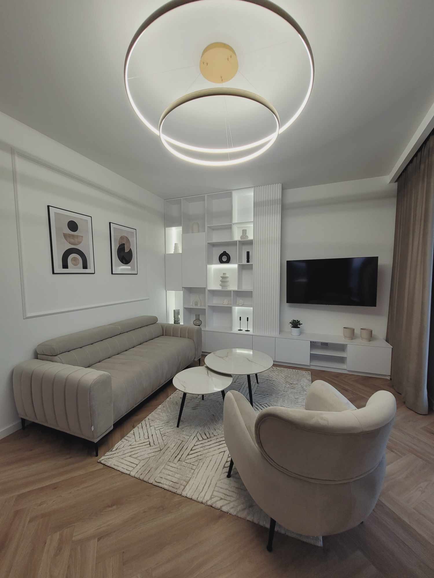 Jasne wnętrze salonu z beżową sofą, fotelami i dywanem w geometryczne wzory. Na ścianie grafiki i regał z podświetleniem. Okrągła lampa sufitowa.