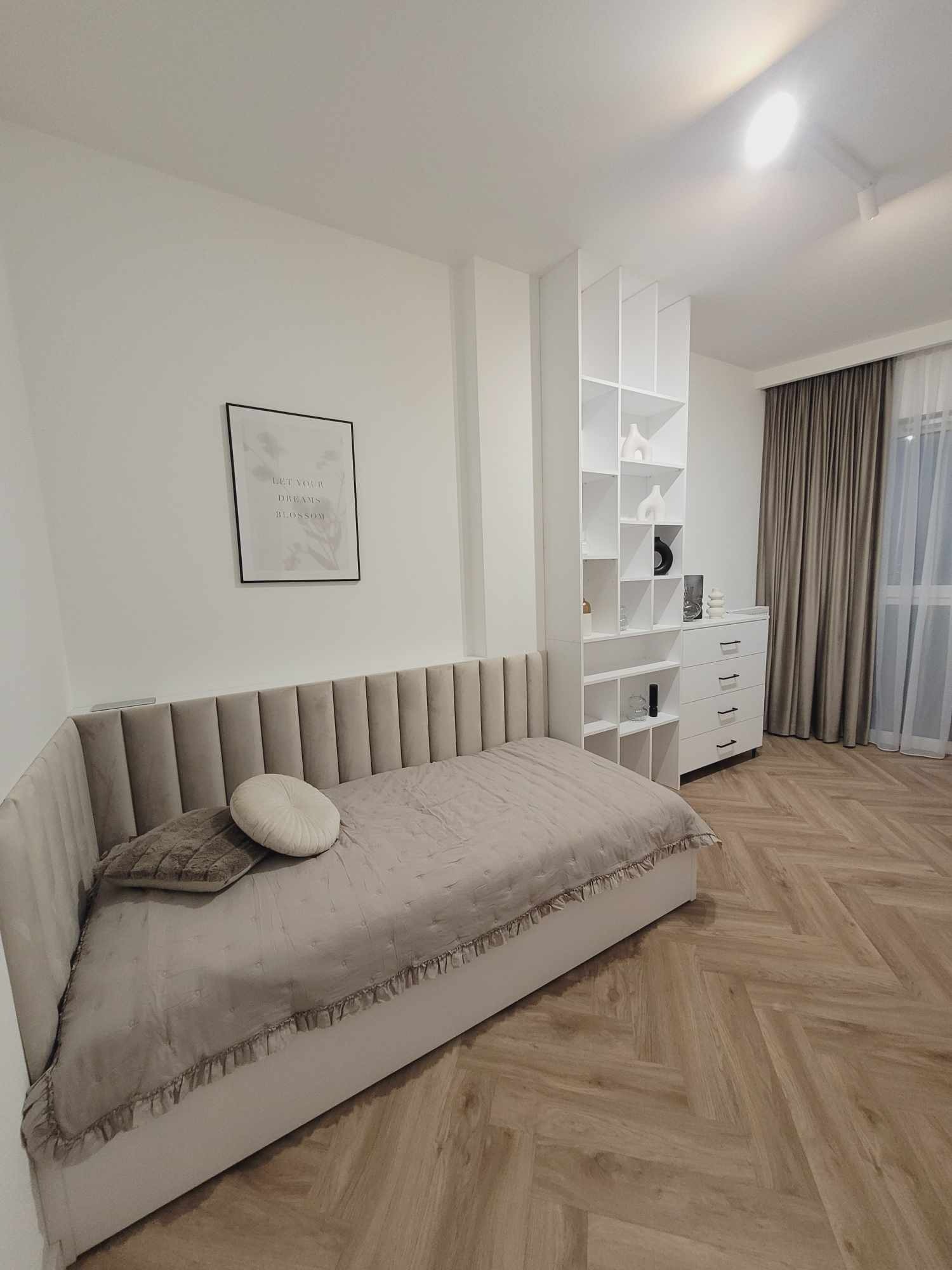 Jasna sypialnia z łóżkiem z wezgłowiem, regałem i komodą. Parkiet ułożony w jodełkę. Na ścianie grafika w czarnej ramie. Stonowana kolorystyka.