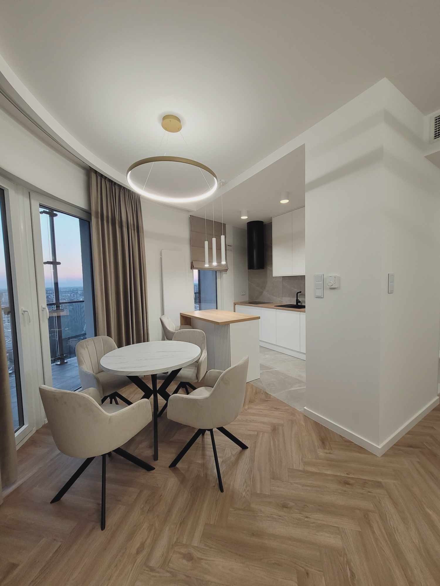 Jasne, nowoczesne wnętrze mieszkania z okrągłym stołem, krzesłami i widokiem na miasto zza okna. Parkiet ułożony w jodełkę. Minimalistyczna kuchnia w tle.