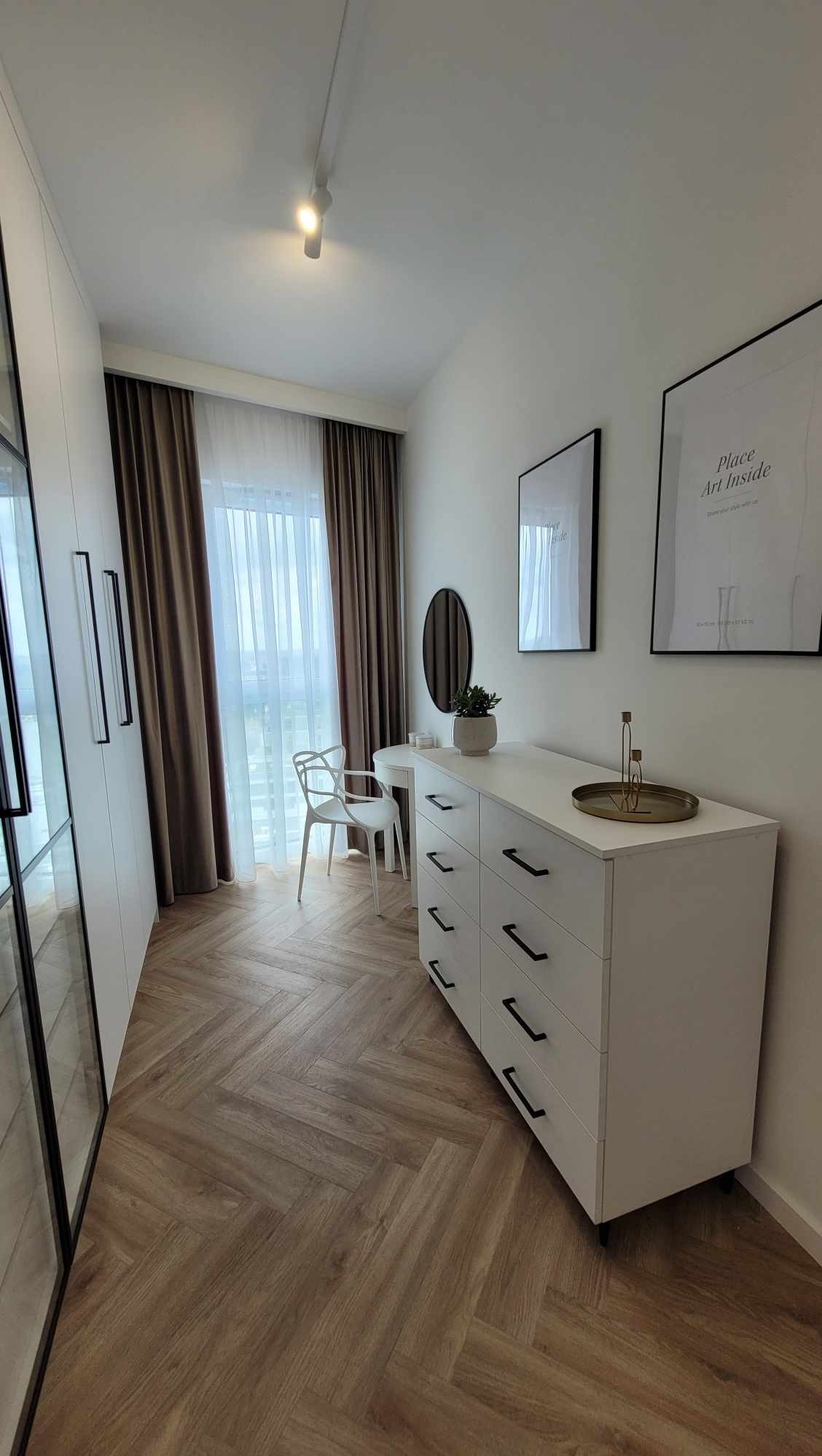 Jasne wnętrze garderoby z komodą, toaletką i lustrem. Parkiet w jodełkę, minimalistyczne dekoracje i duże okno z brązowymi zasłonami. Nowoczesny design.