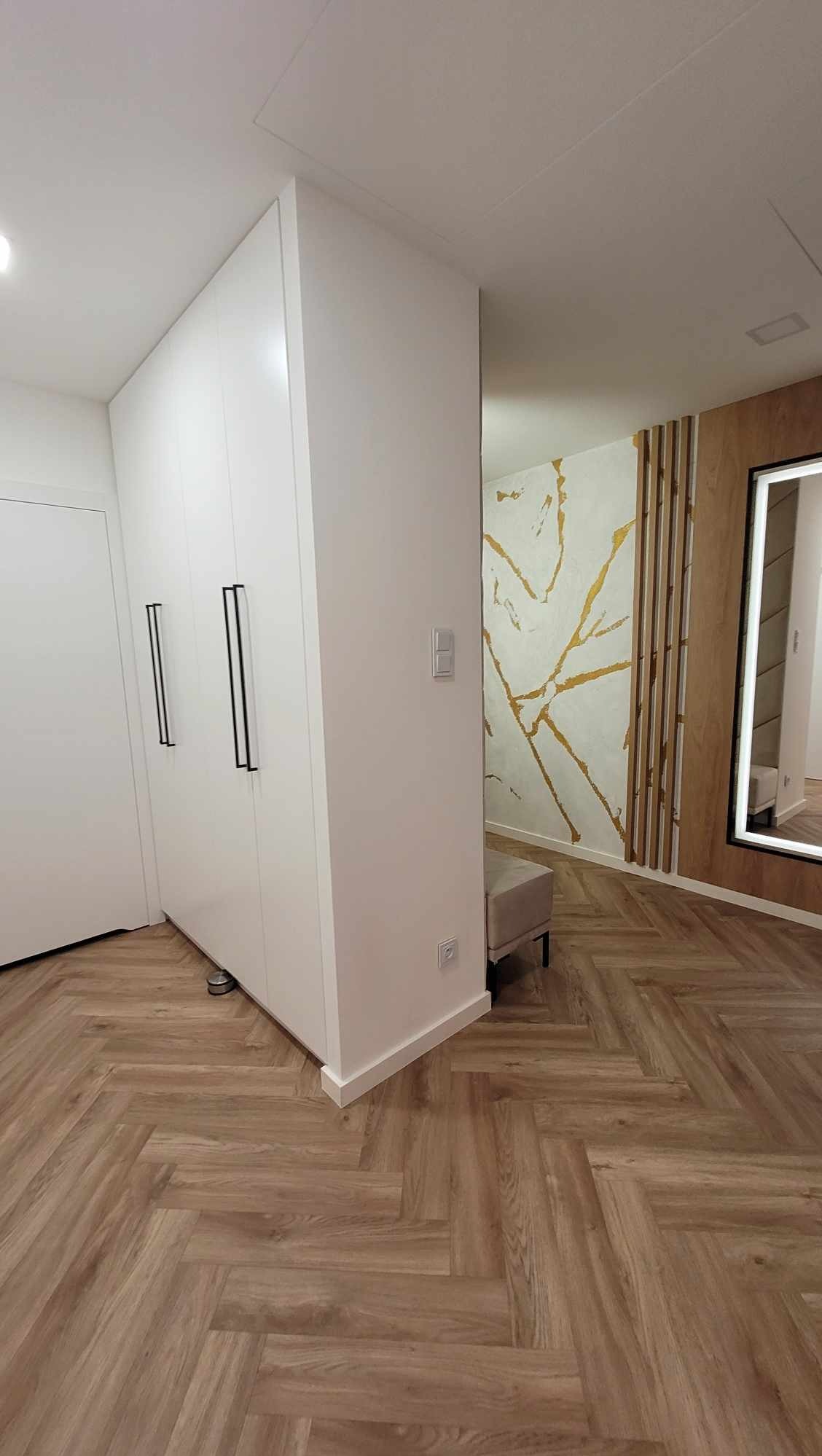 Biała szafa z czarnymi uchwytami w przedpokoju z parkietem w jodełkę. Na ścianie dekoracyjna tapeta z motywem złotych żył. Lustro z podświetleniem.