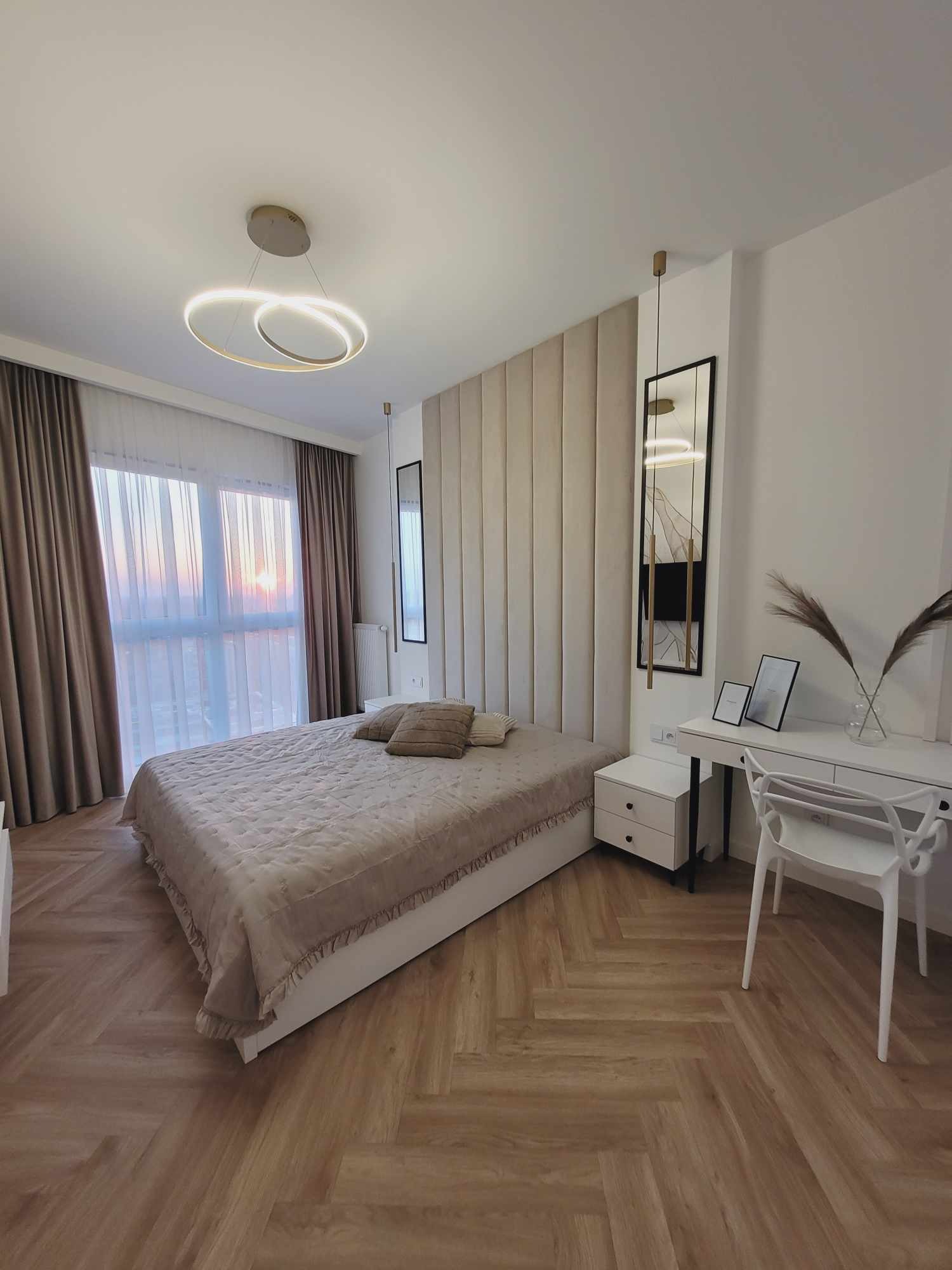 Jasna sypialnia z łóżkiem, panelem tapicerowanym za wezgłowiem i toaletką z krzesłem. Uwagę przykuwa nowoczesna lampa wisząca i parkiet ułożony w jodełkę.