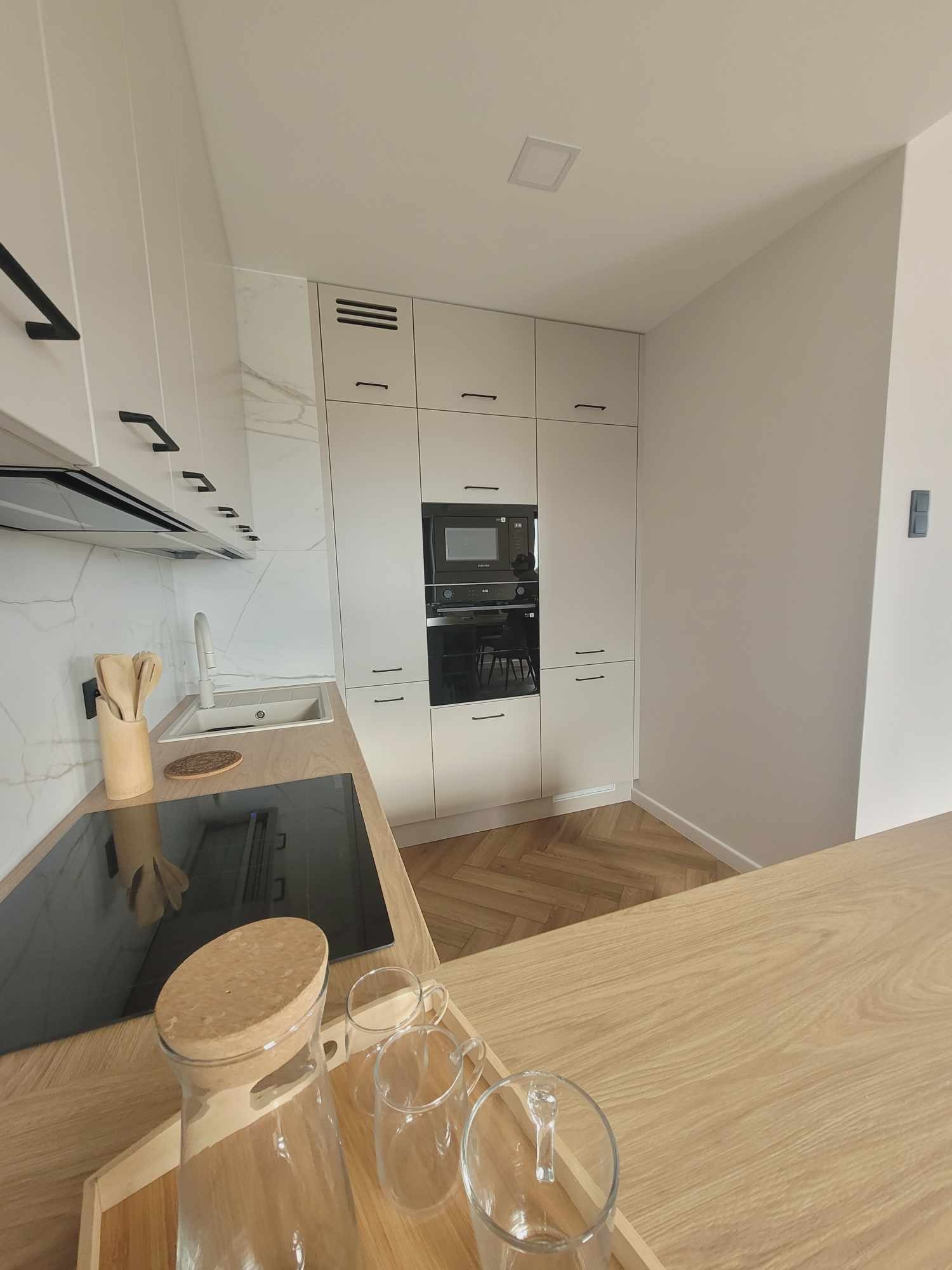 Jasna kuchnia z drewnianym blatem i podłogą w jodełkę. Białe szafki z czarnymi uchwytami, czarna płyta indukcyjna i sprzęt AGD. Na blacie szklane naczynia.