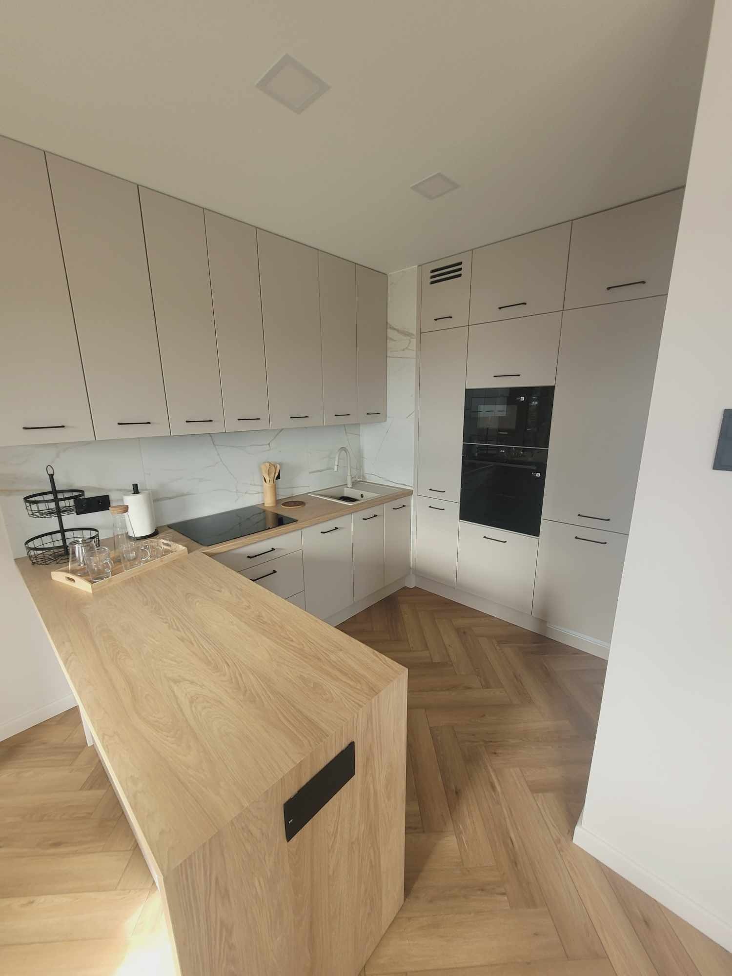 Jasna kuchnia z drewnianym blatem i parkietem w jodełkę. Minimalistyczne szafki z czarnymi uchwytami. Na blacie akcesoria kuchenne i szklanki na tacy.