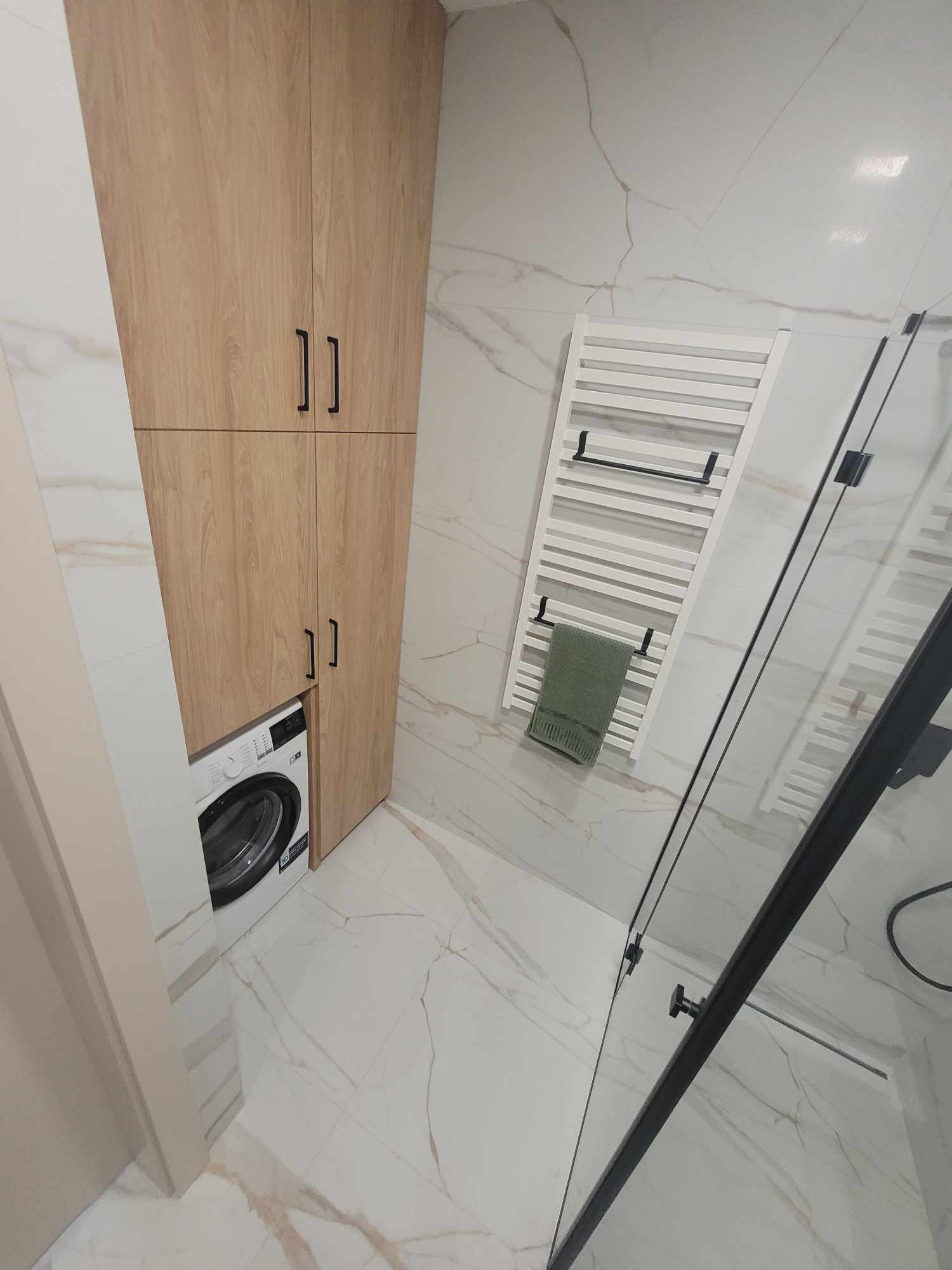 Jasna łazienka z drewnianą szafą zabudowaną nad pralką, białe marmurowe płytki, grzejnik drabinkowy z ręcznikiem i kabina prysznicowa z czarnymi profilami.