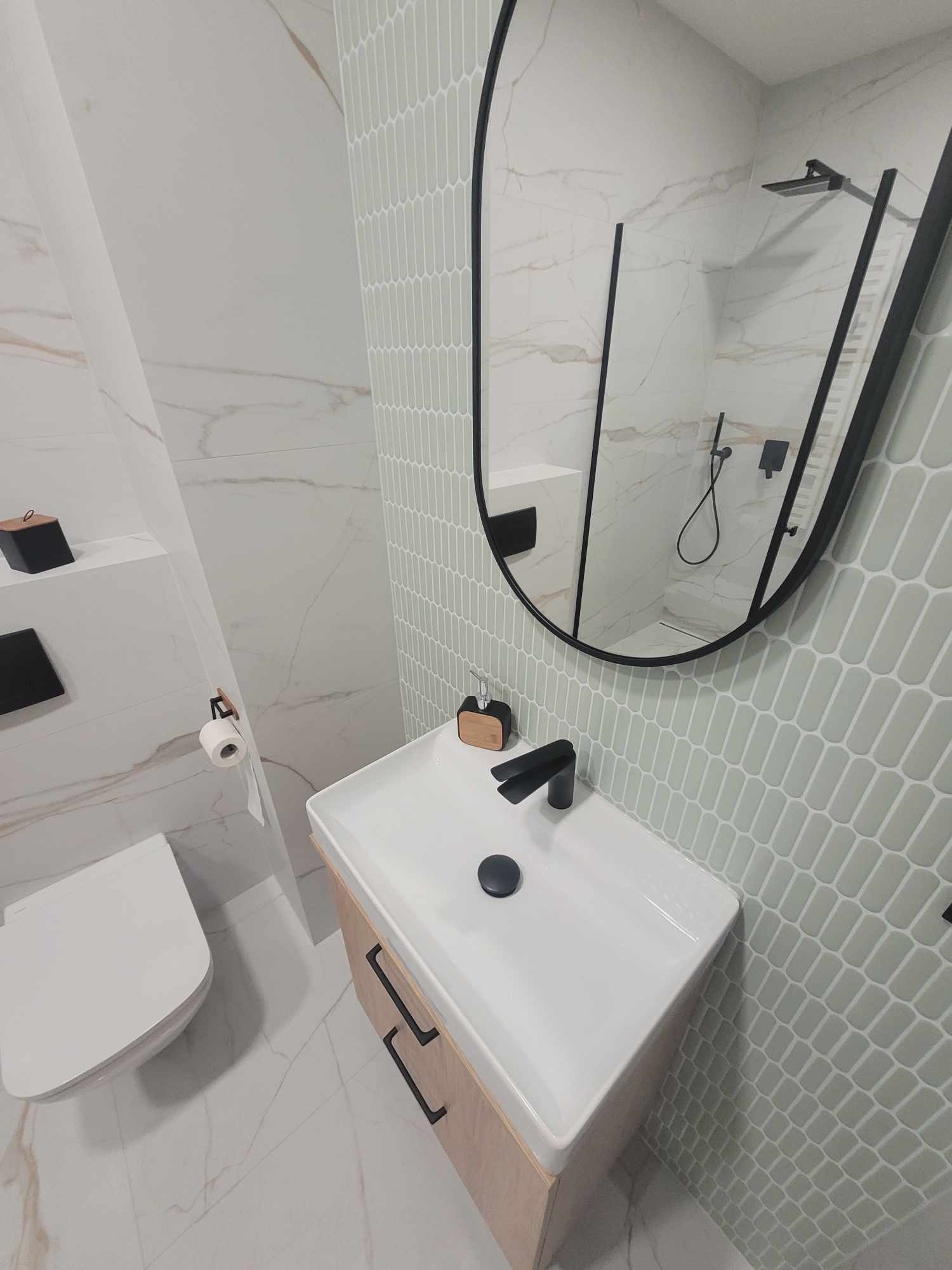 Nowoczesna łazienka z umywalką nablatową, czarną armaturą i lustrem w czarnej ramie. Białe płytki imitujące marmur i mozaika w kształcie pastylek.