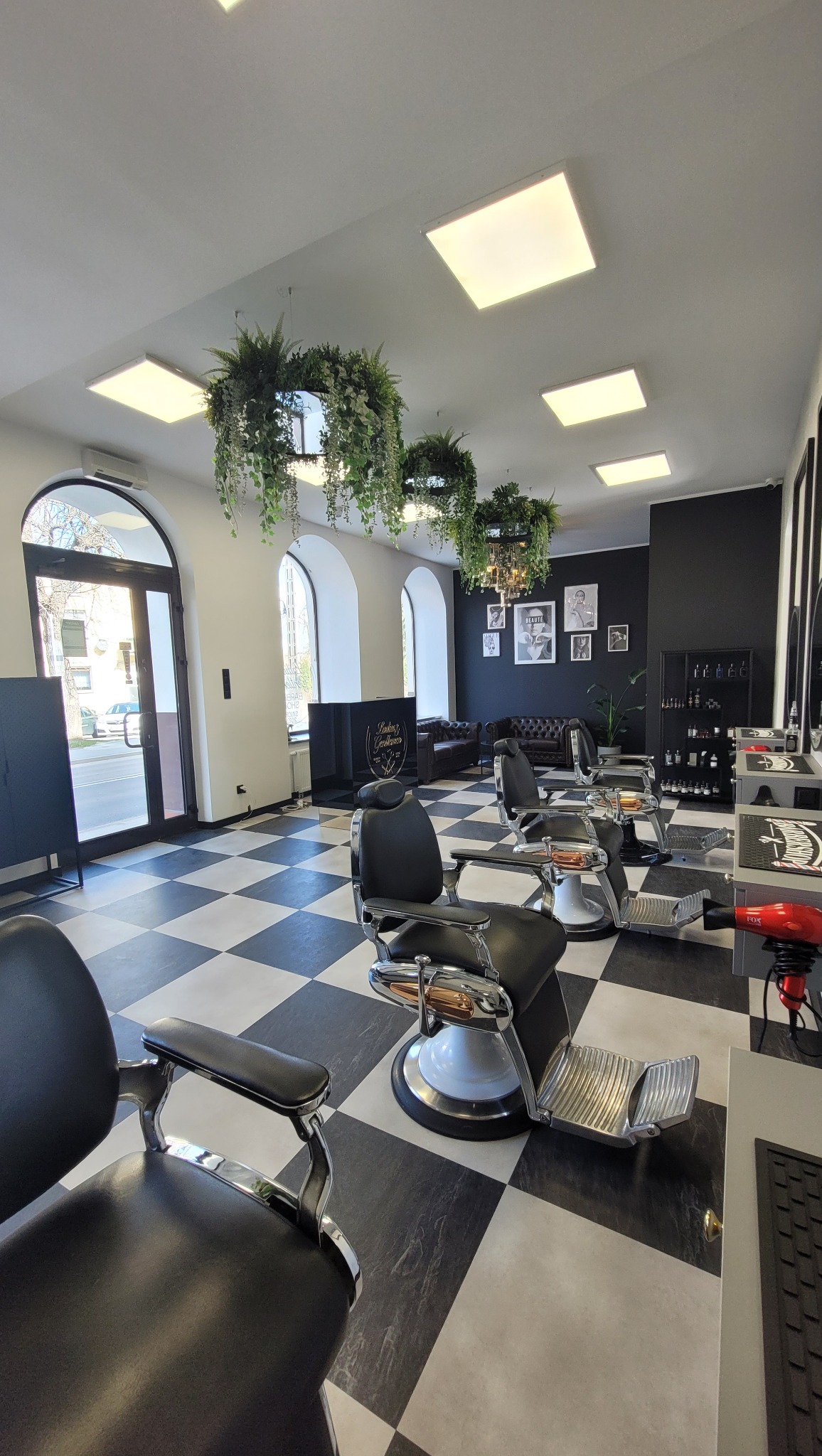 Nowoczesne wnętrze salonu fryzjerskiego z czarno-białą szachownicą na podłodze, roślinami pod sufitem i fotelami fryzjerskimi w stylu retro.