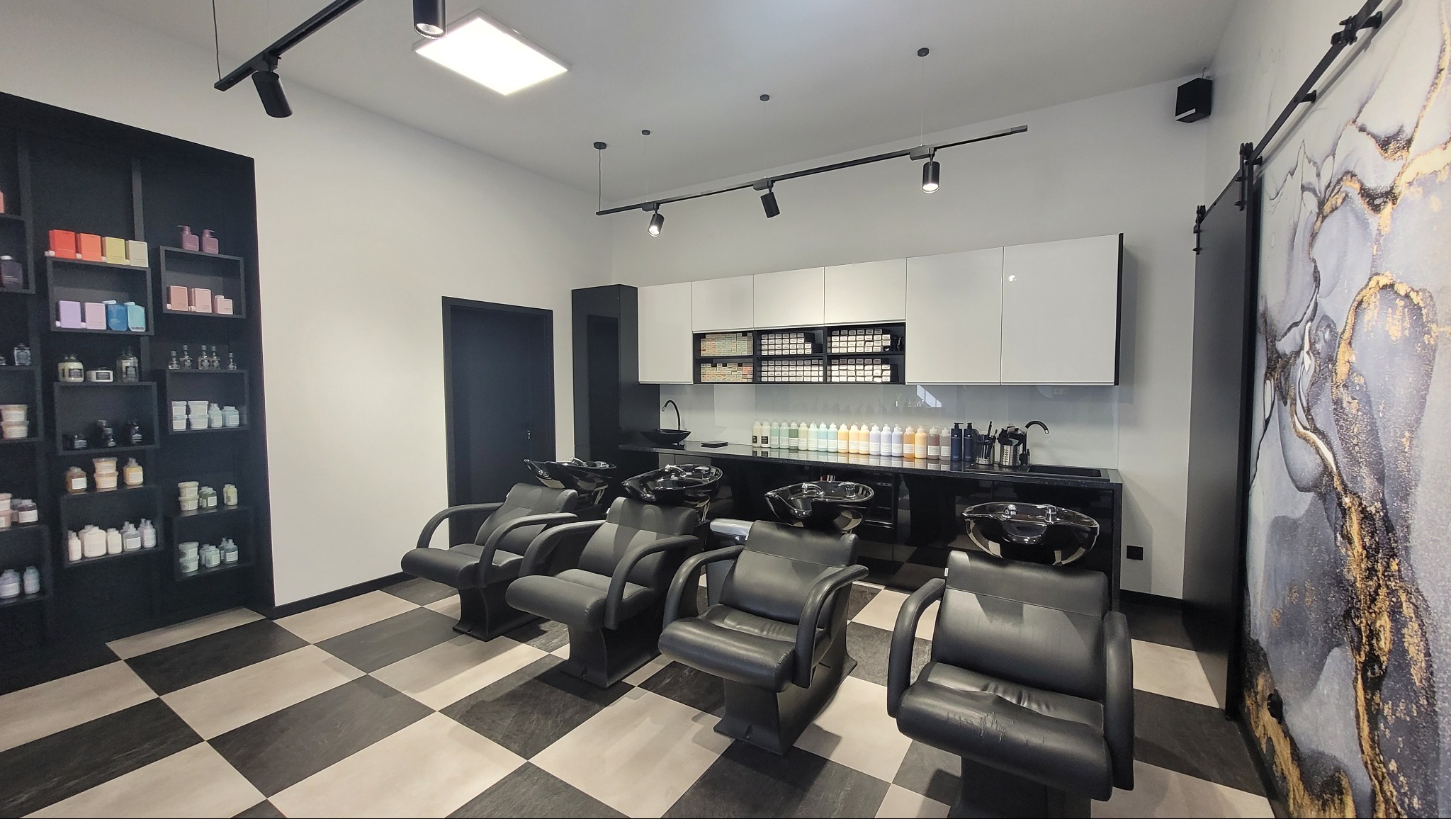 Nowoczesne wnętrze salonu fryzjerskiego z czterema stanowiskami, czarno-białą szachownicą na podłodze i minimalistycznym wystrojem. Widoczne kosmetyki i akcesoria.