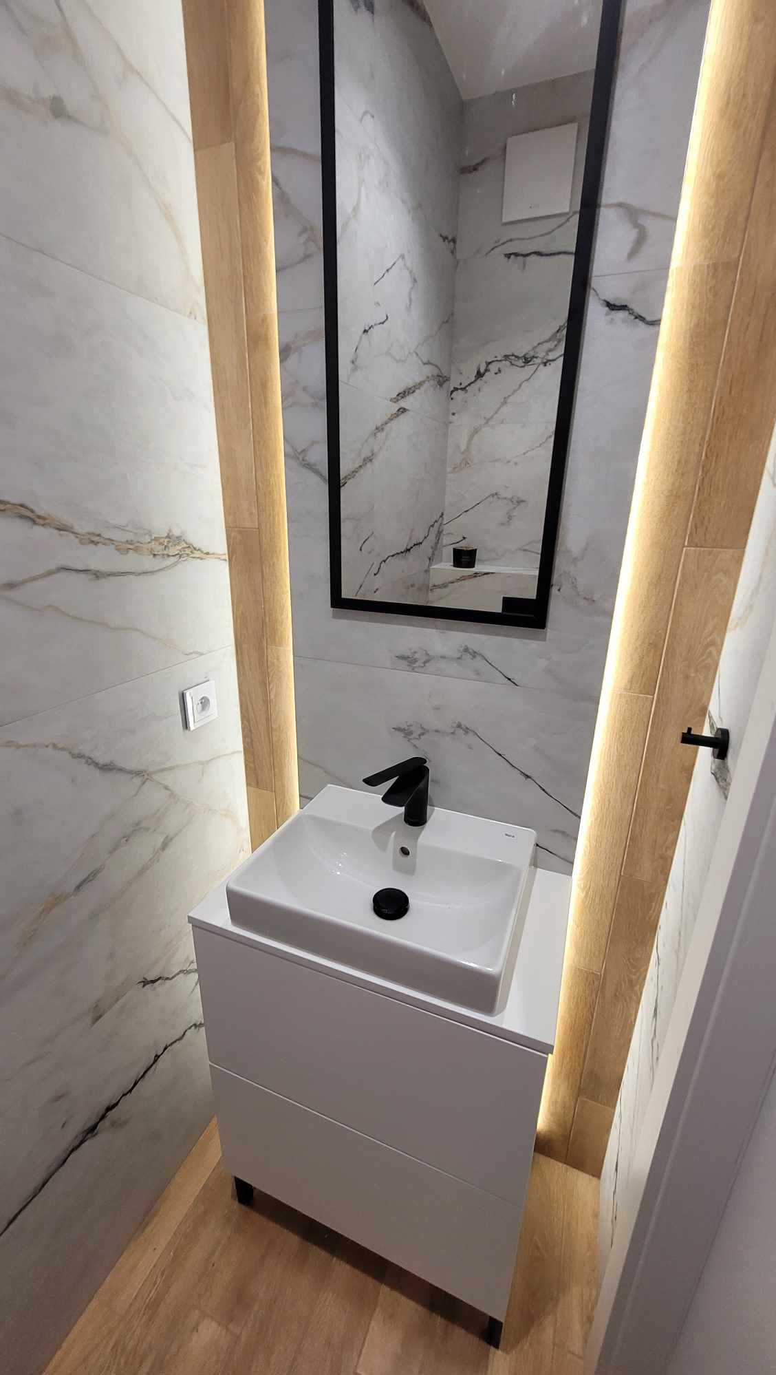 Nowoczesna łazienka z umywalką nablatową, czarną armaturą i lustrem w czarnej ramie. Marmurowe płytki ścienne i podświetlane drewniane panele.