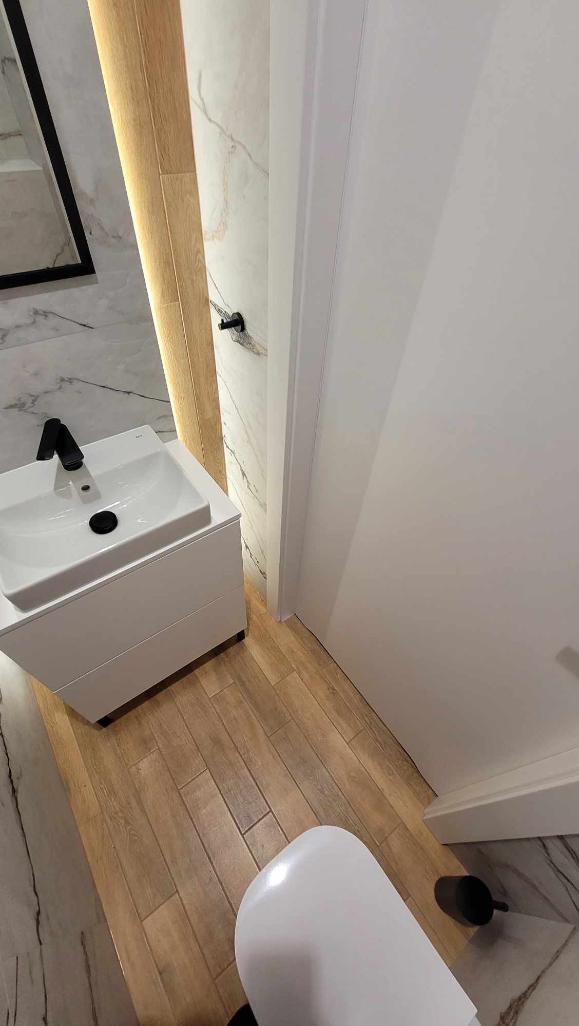 Nowoczesna łazienka z białą umywalką nablatową i armaturą w kolorze czarnym. Marmurowe płytki ścienne i drewnopodobna podłoga tworzą elegancki kontrast.