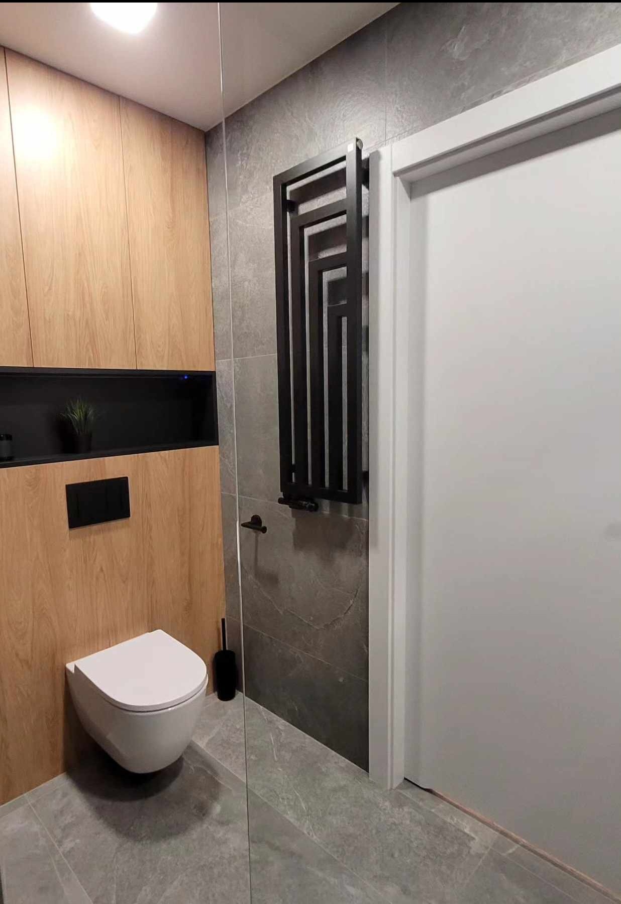 Nowoczesna łazienka z drewnianą zabudową, szarą płytką i czarnym grzejnikiem drabinkowym. Wisząca toaleta i minimalistyczne dodatki.