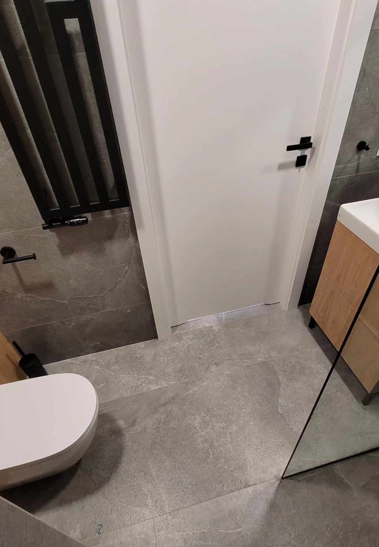 Nowoczesna łazienka z szarymi płytkami, białą armaturą i czarnymi akcentami: grzejnik drabinkowy, bateria umywalkowa i klamka. Minimalistyczny design.