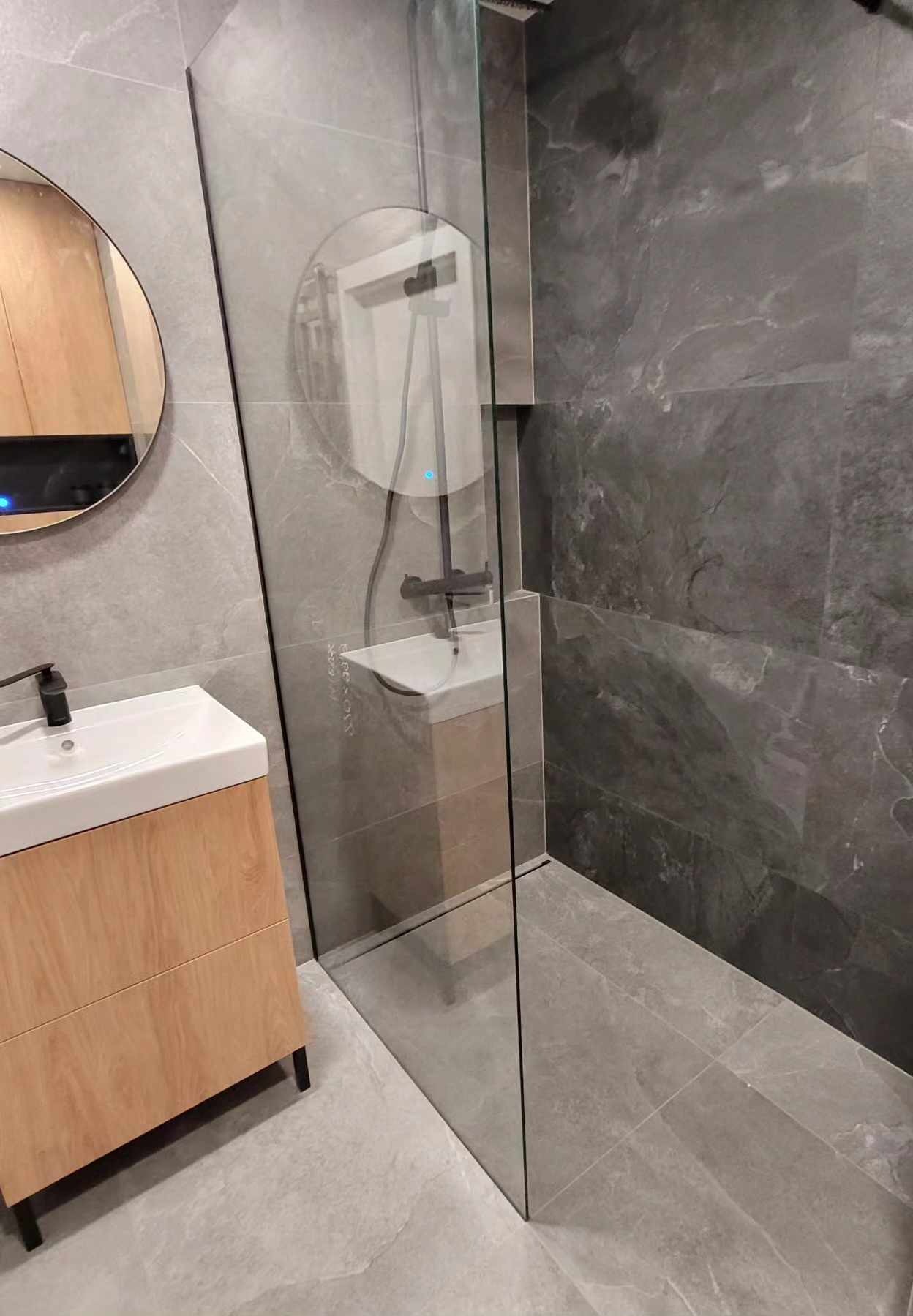 Nowoczesna łazienka z kabiną typu walk-in, umywalką na szafce z drewnianym frontem i okrągłym lustrem. Szare płytki imitujące kamień na ścianach i podłodze.