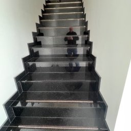 Orpol Sp.z o.o. - Polerowane, czarne schody kamienne z subtelnymi, metalowymi akcentami, widziane z dołu. W lustrzanej powierzchni odbija się sylwetka fotografa.