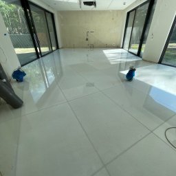 Orpol Sp.z o.o. - Jasne, kwadratowe płyty tarasowe w przestronnym wnętrzu z dużymi oknami. Widoczne przygotowania do dalszych prac wykończeniowych, kable i rury.