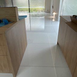 Orpol Sp.z o.o. - Błyszcząca, biała podłoga z płytek w nowym wnętrzu. Widoczne fragmenty zabudowy meblowej z jasnego drewna. Duże okno z widokiem na zieleń. Minimalistyczny design.