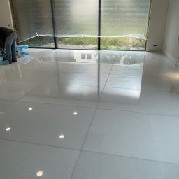 Orpol Sp.z o.o. - Błyszcząca, jasna posadzka z płytek w trakcie montażu. Widoczny pracownik docinający płytki przy dużym oknie. Podłoga zabezpieczona folią w tle.