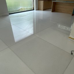 Orpol Sp.z o.o. - Błyszcząca, jasna podłoga z kamiennych płyt w kwadratowym wzorze, odbijająca światło z okna. Widoczne fragmenty mebli kuchennych w tle.