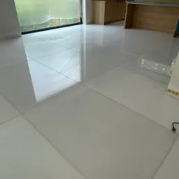 Błyszcząca, jasna podłoga z kamiennych płyt w kwadratowym wzorze, odbijająca światło z okna. Widoczne fragmenty mebli kuchennych w tle.