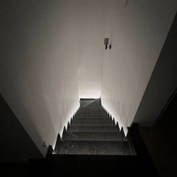 Orpol Sp.z o.o. - Nowoczesne schody kamienne z podświetleniem LED, widok z góry w dół. Minimalistyczny design, ciemne stopnie, jasne ściany i sufit. Oświetlenie punktowe.