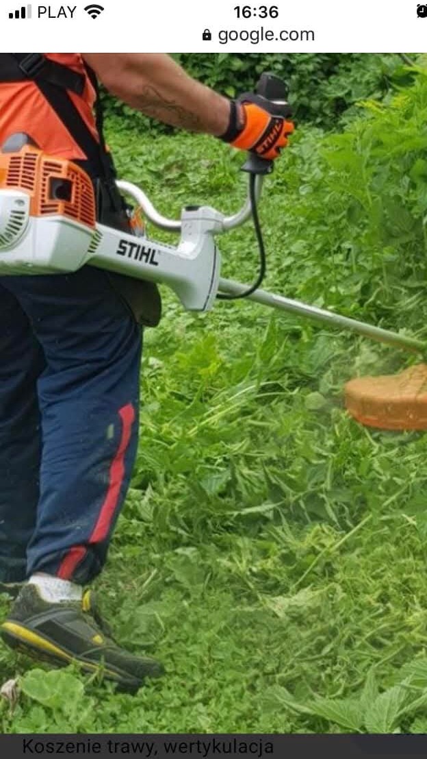 Pracownik w pomarańczowej koszulce kosi trawę kosiarką spalinową Stihl. Widoczne skoszone źdźbła trawy i wysoka zieleń w tle. Ujęcie z boku, na pierwszym planie buty.