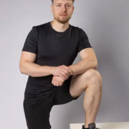 Młody mężczyzna w czarnym sportowym stroju, oparty nogą o biały sześcian, pozuje na szarym tle. Widoczny łańcuszek na szyi i szare buty sportowe.