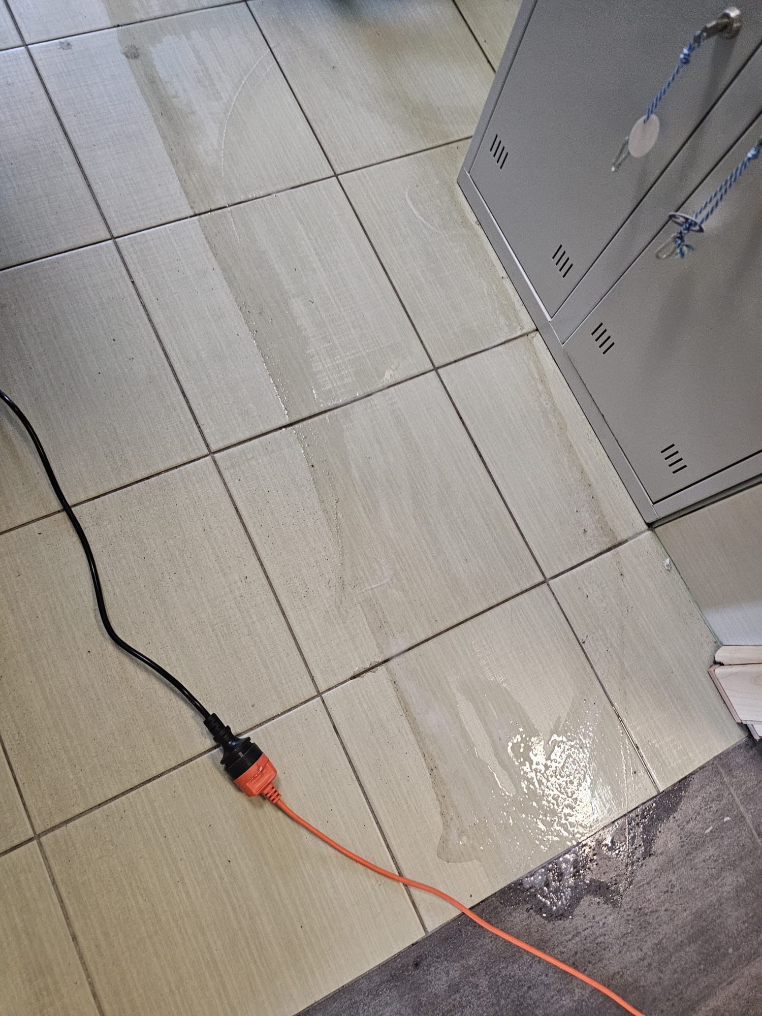 Czyszczona podłoga z płytek, widoczne mokre ślady po myciu. W rogu metalowa szafka i kabel zasilający. Ujęcie z góry, skupione na procesie czyszczenia.