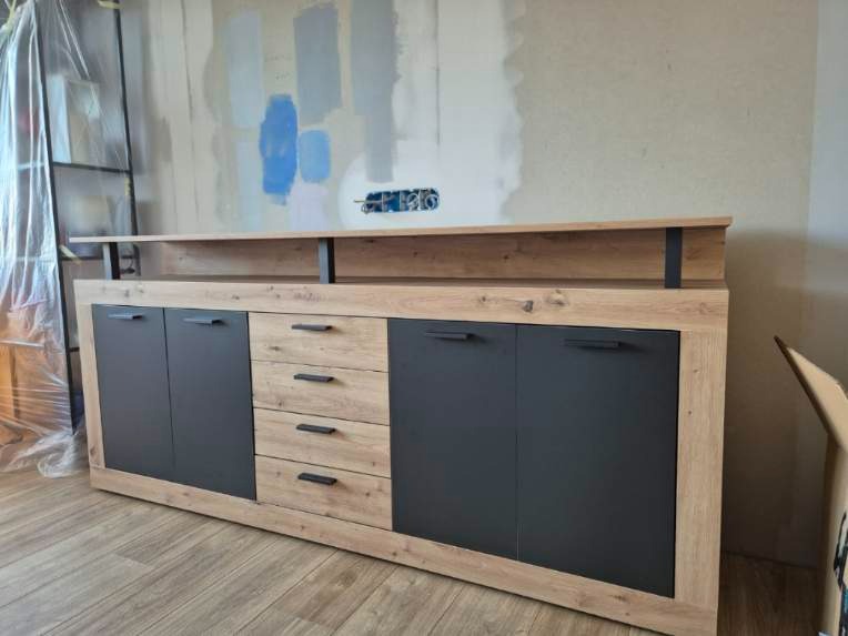 Zmontowana komoda z szufladami i szafkami w odcieniach drewna i grafitu, na tle ściany z widocznymi śladami po malowaniu i regału zabezpieczonego folią.