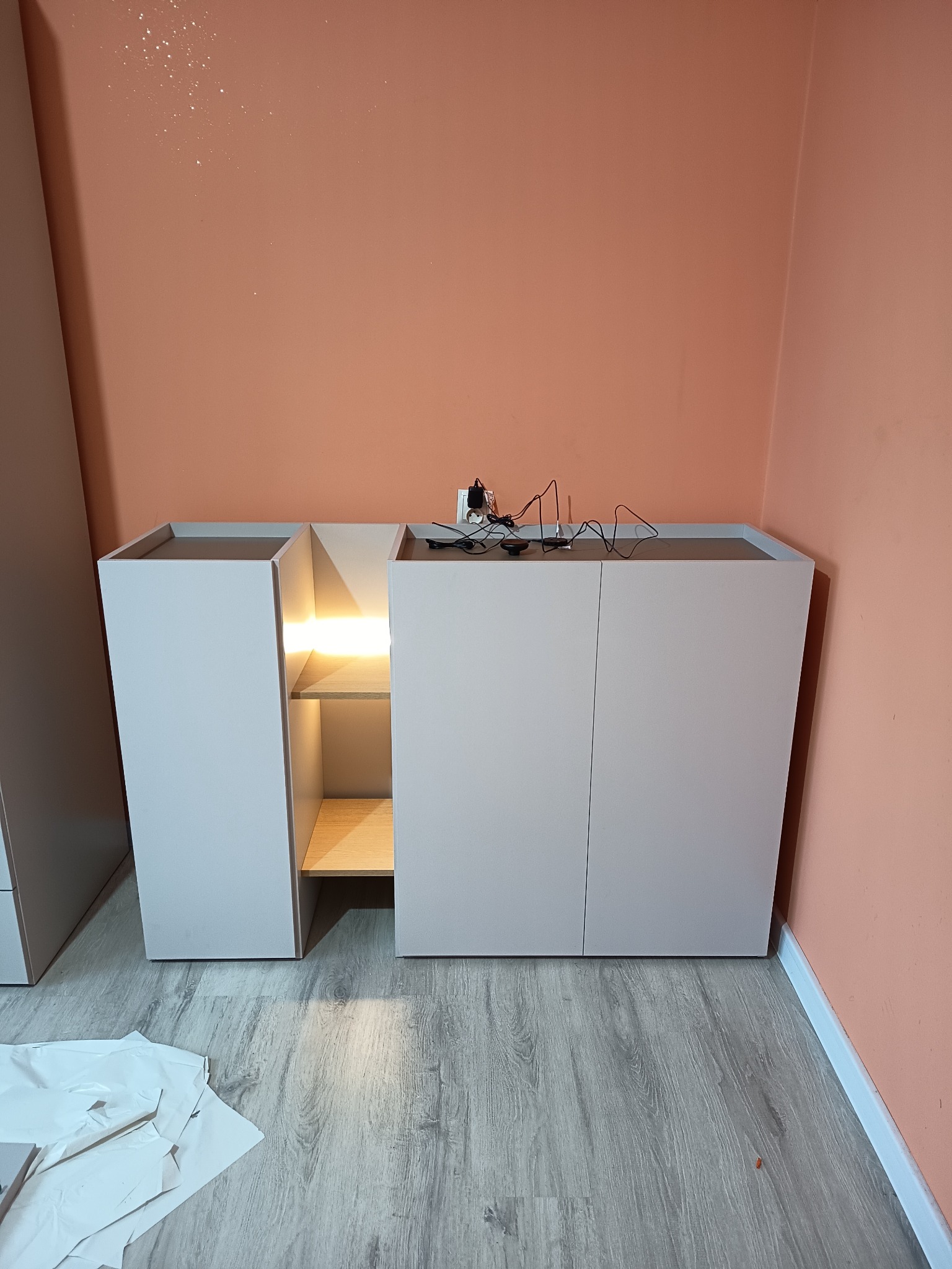 Nowoczesna komoda z oświetleniem LED po montażu w Bielsku Podlaskim. Widoczne kable na blacie, szare panele podłogowe i pomarańczowa ściana w tle.