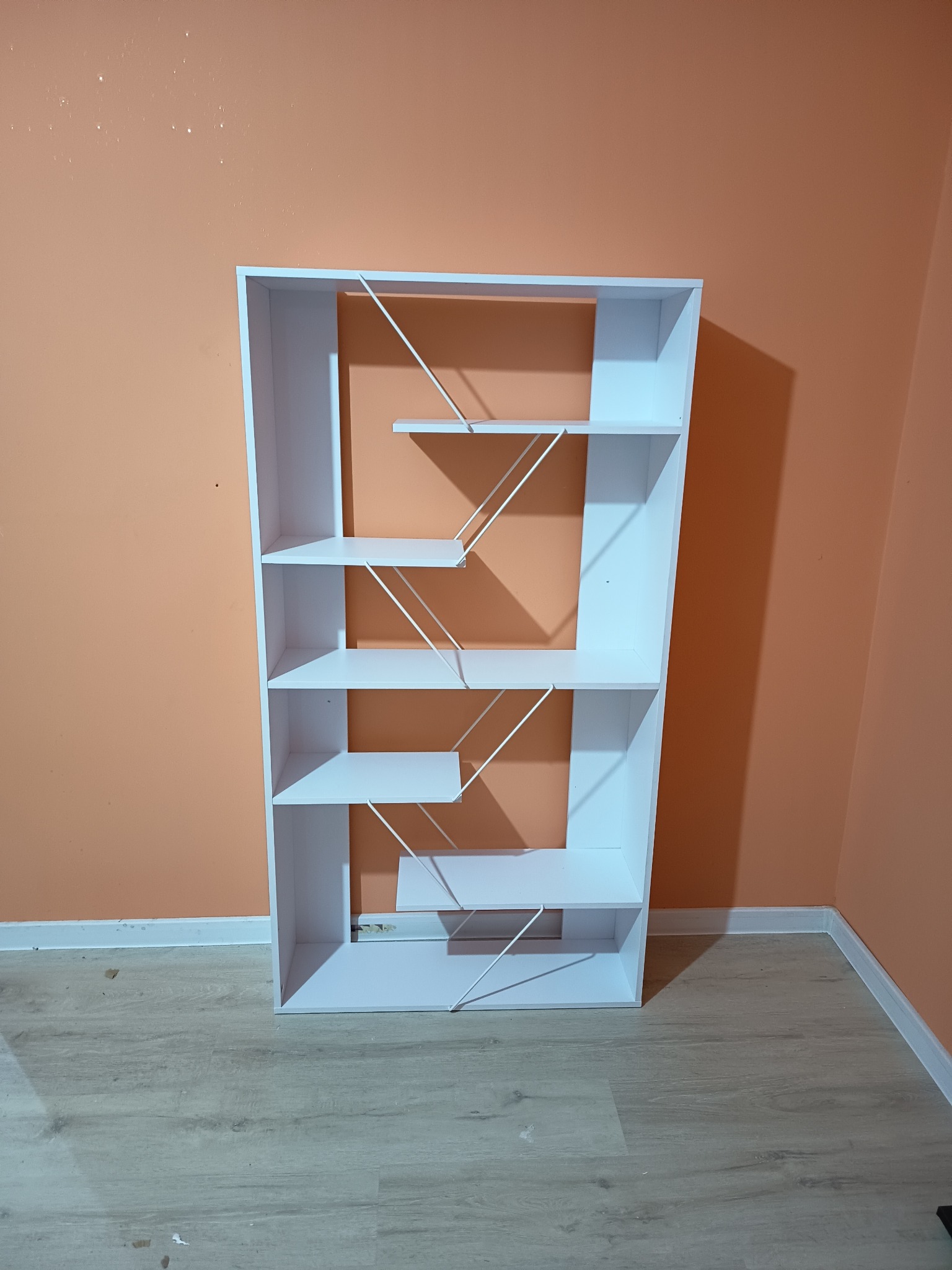 Biały, minimalistyczny regał z asymetrycznymi półkami na tle brzoskwiniowej ściany. Widoczne detale konstrukcji i jasna podłoga.