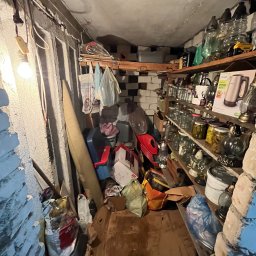 Robimy Robotę ROBERT BALCERAK - Zagracone pomieszczenie w Głownie: półki z słoikami i różnymi przedmiotami, kartony i worki na podłodze, surowa ściana z cegły i wisząca żarówka.