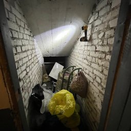 Robimy Robotę ROBERT BALCERAK - Zakamarek ze ścianami z białej cegły wypełniony workami i odpadami, oświetlony lampą roboczą. Widoczne narzędzie w tle. Nierówna podłoga i sufit.