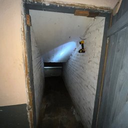 Robimy Robotę ROBERT BALCERAK - Wejście do piwnicy z oświetleniem roboczym przymocowanym do ściany. Widoczne białe cegły i drewniane elementy konstrukcyjne. Perspektywa zagłębiająca się w ciemność.