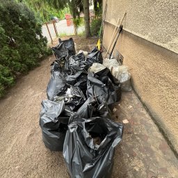 Robimy Robotę ROBERT BALCERAK - Stos czarnych worków na śmieci z liśćmi i gałęziami obok ściany budynku i ogrodzenia. Grabie i inne narzędzia opierają się o ścianę.