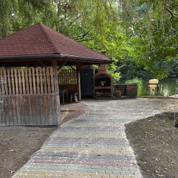 Robimy Robotę ROBERT BALCERAK - Kolorowa kostka brukowa prowadzi do drewnianej altany z grillem, usytuowanej nad brzegiem wody, otoczonej zielenią drzew.