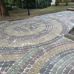 Robimy Robotę ROBERT BALCERAK - Kolorowa kostka brukowa ułożona w spiralny wzór na podjeździe. Widok na dom i zieleń w tle. Artystyczne ułożenie kostki brukowej w ogrodzie.