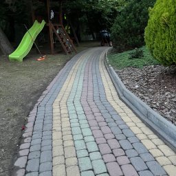 Robimy Robotę ROBERT BALCERAK - Kolorowa kostka brukowa tworząca ścieżkę w ogrodzie, prowadząca obok trawnika i zieleni, z placem zabaw w tle. Widoczne narzędzie do zamiatania.