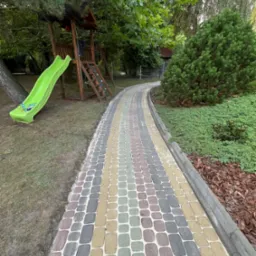 Kolorowa kostka brukowa tworzy krętą ścieżkę w ogrodzie z placem zabaw, zjeżdżalnią i bujną zielenią. Krawężnik oddziela ścieżkę od rabat z korą.