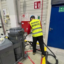 Robimy Robotę ROBERT BALCERAK - Pracownik w kamizelce z napisem 'ROBIMY ROBOTĘ' obsługuje przemysłowy odkurzacz w hali. Widoczny sprzęt ppoż. i oznakowania. Hala z betonową podłogą.