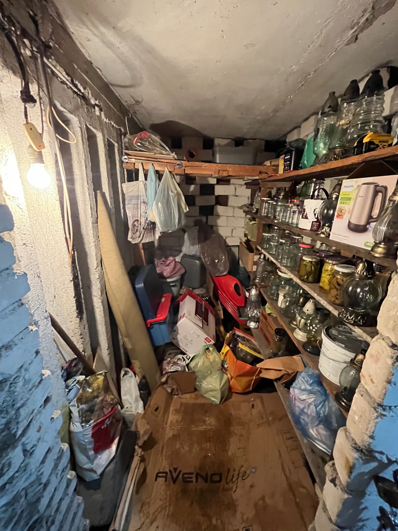 Zagracone pomieszczenie w Głownie: półki z słoikami i różnymi przedmiotami, kartony i worki na podłodze, surowa ściana z cegły i wisząca żarówka.