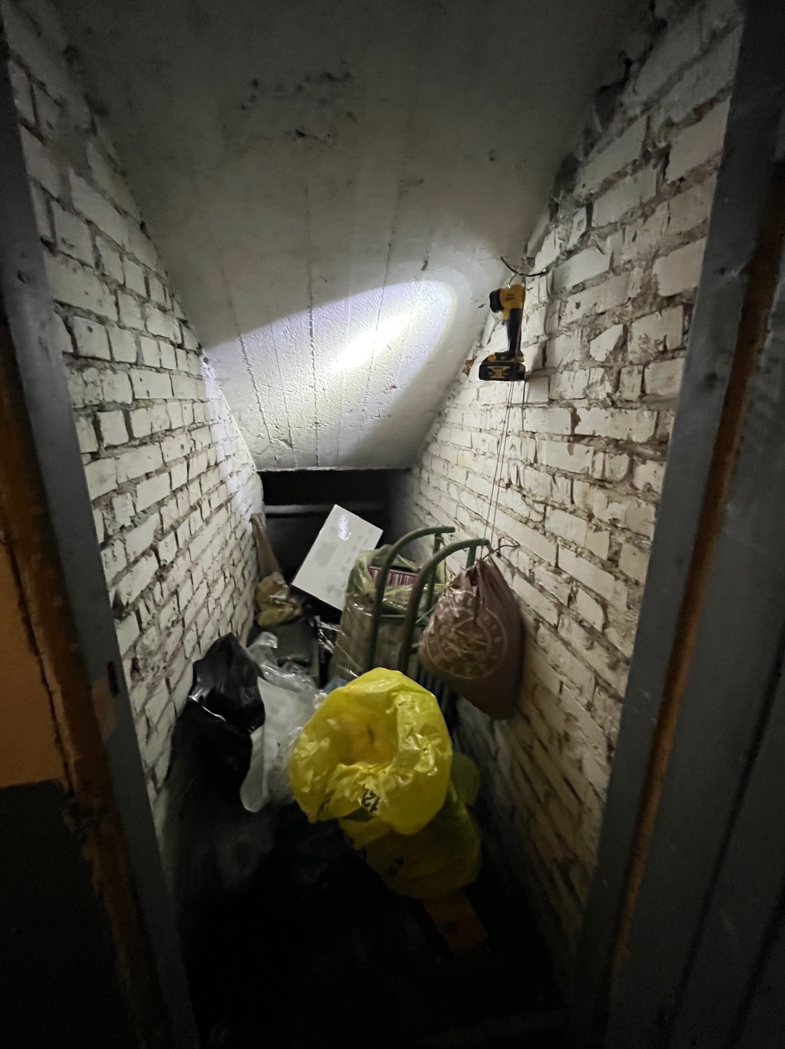 Zakamarek ze ścianami z białej cegły wypełniony workami i odpadami, oświetlony lampą roboczą. Widoczne narzędzie w tle. Nierówna podłoga i sufit.