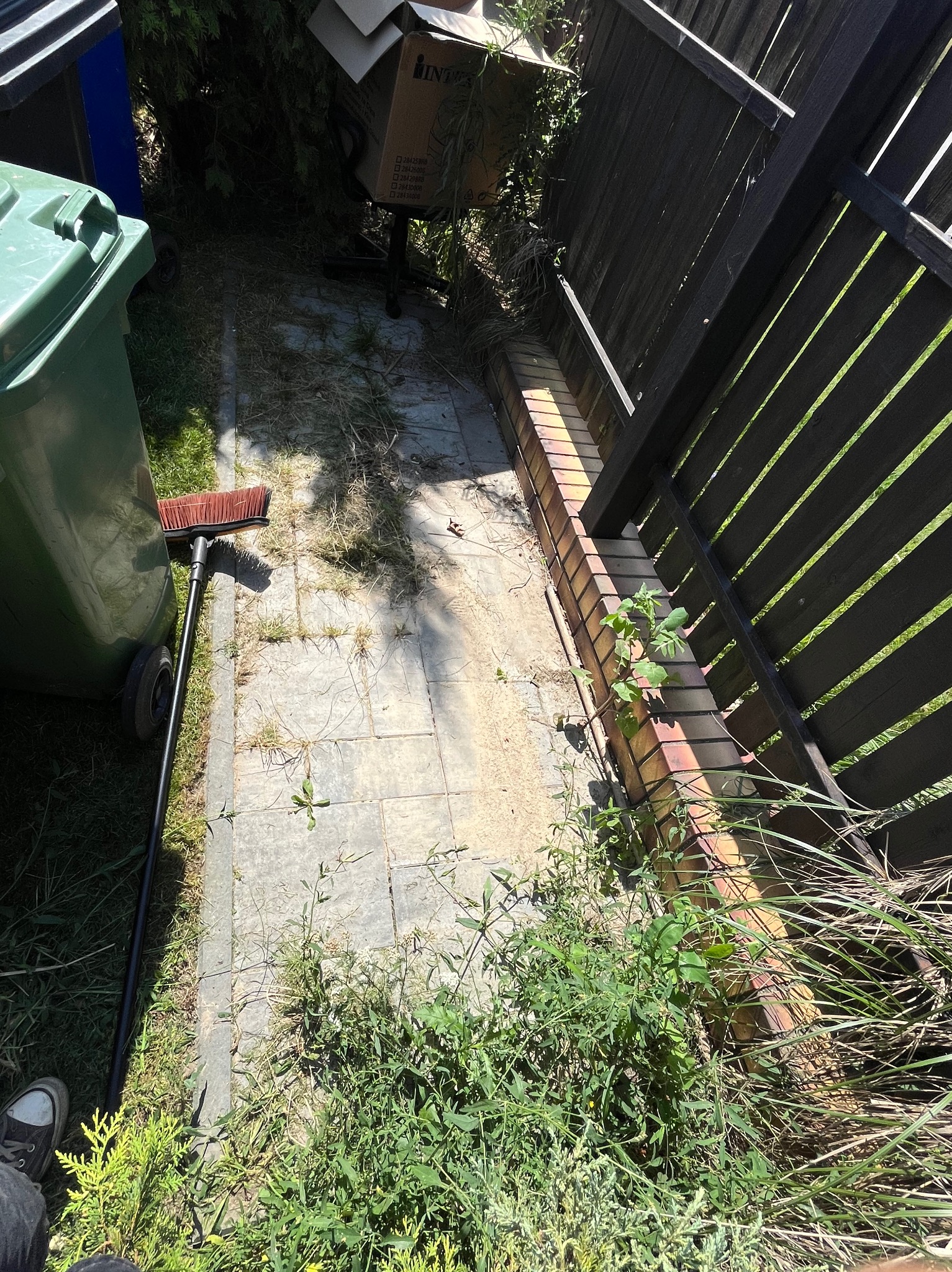 Zaniedbana kostka brukowa z chwastami, liśćmi i piaskiem. Widoczny kosz na śmieci, szczotka i fragment ogrodzenia. Niezadbana przestrzeń.