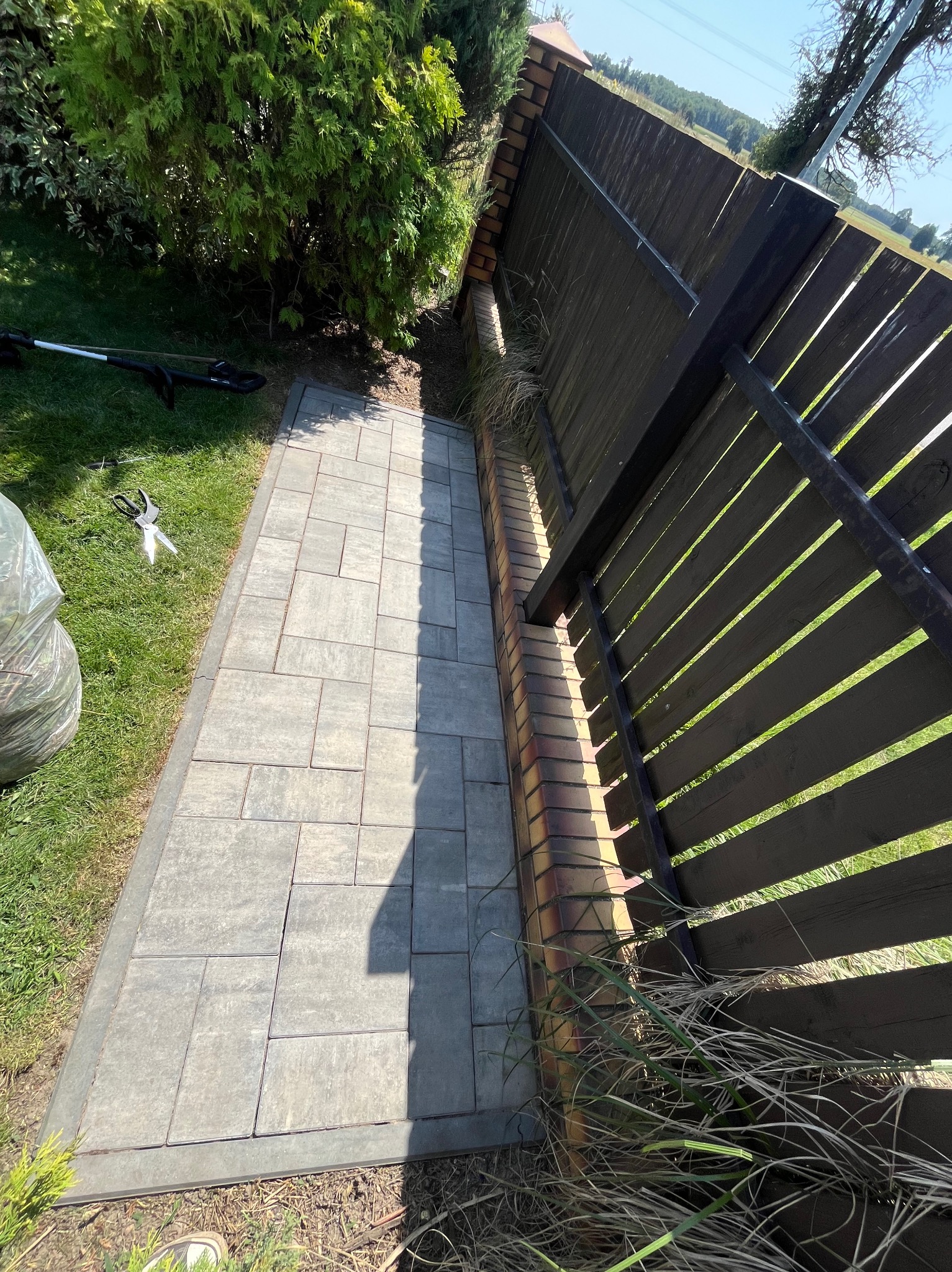 Ułożona kostka brukowa tworząca ścieżkę wzdłuż ogrodzenia. Obok trawa i narzędzia ogrodnicze, sugerujące prace porządkowe. Jasne, słoneczne oświetlenie.